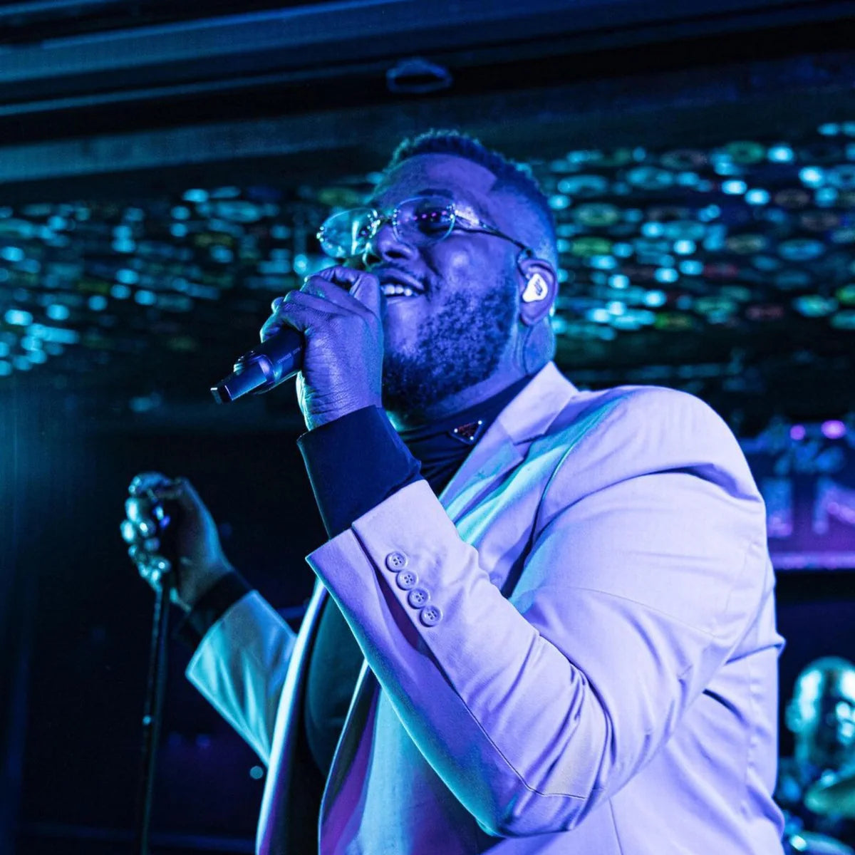 Guordan sings into a handheld microphone, wearing glasses, in-ear monitors and a light jacket under blue stage lights.